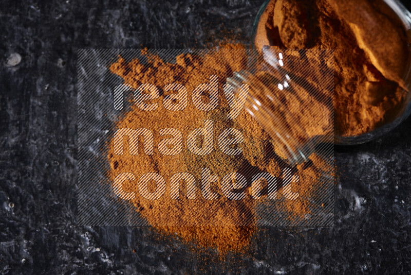 A glass jar full of ground paprika powder flipped with some spilling powder on black background