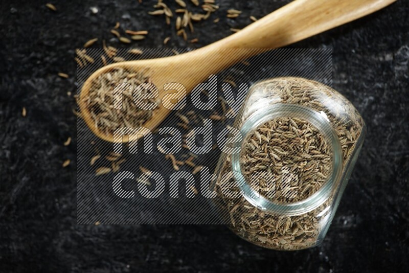 A glass spice jar and a wooden spoon full of cumin seeds on a textured black flooring