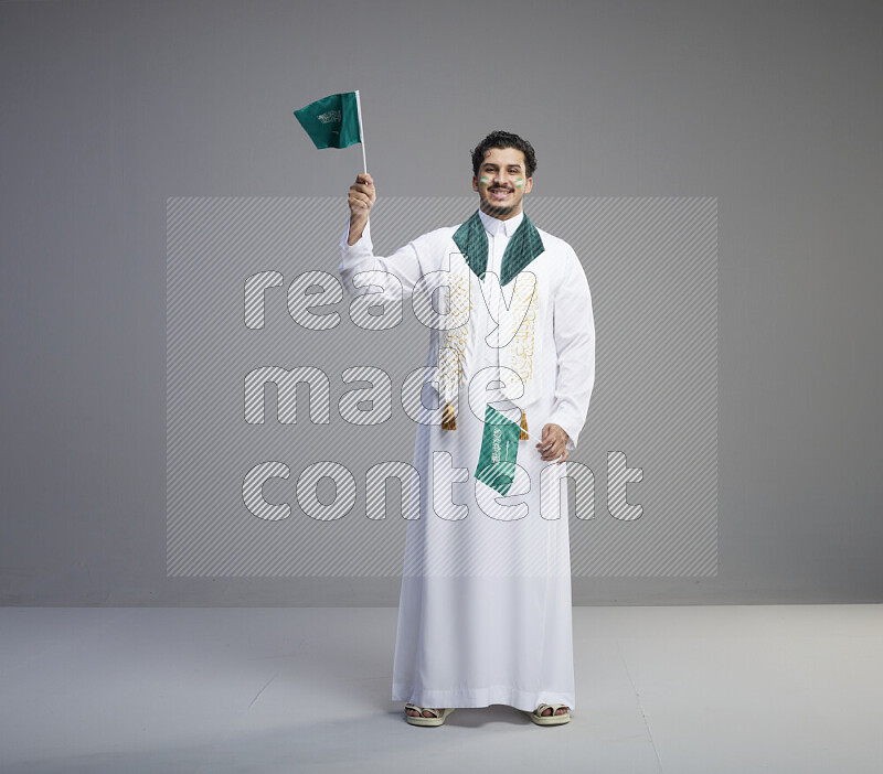 A Saudi man standing wearing thob and saudi flag scarf with face painting holding small Saudi flag on gray background