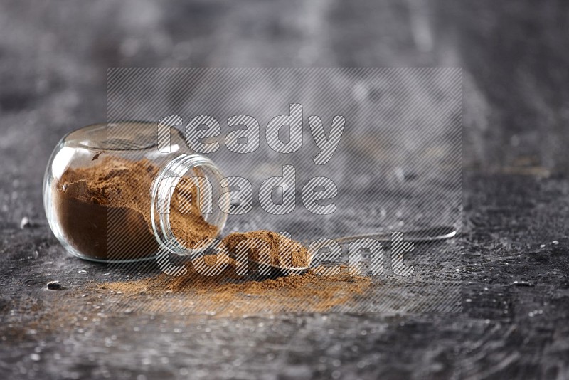 Herbal glass jar full of cinnamon powder flipped and a metal spoon on textured black background