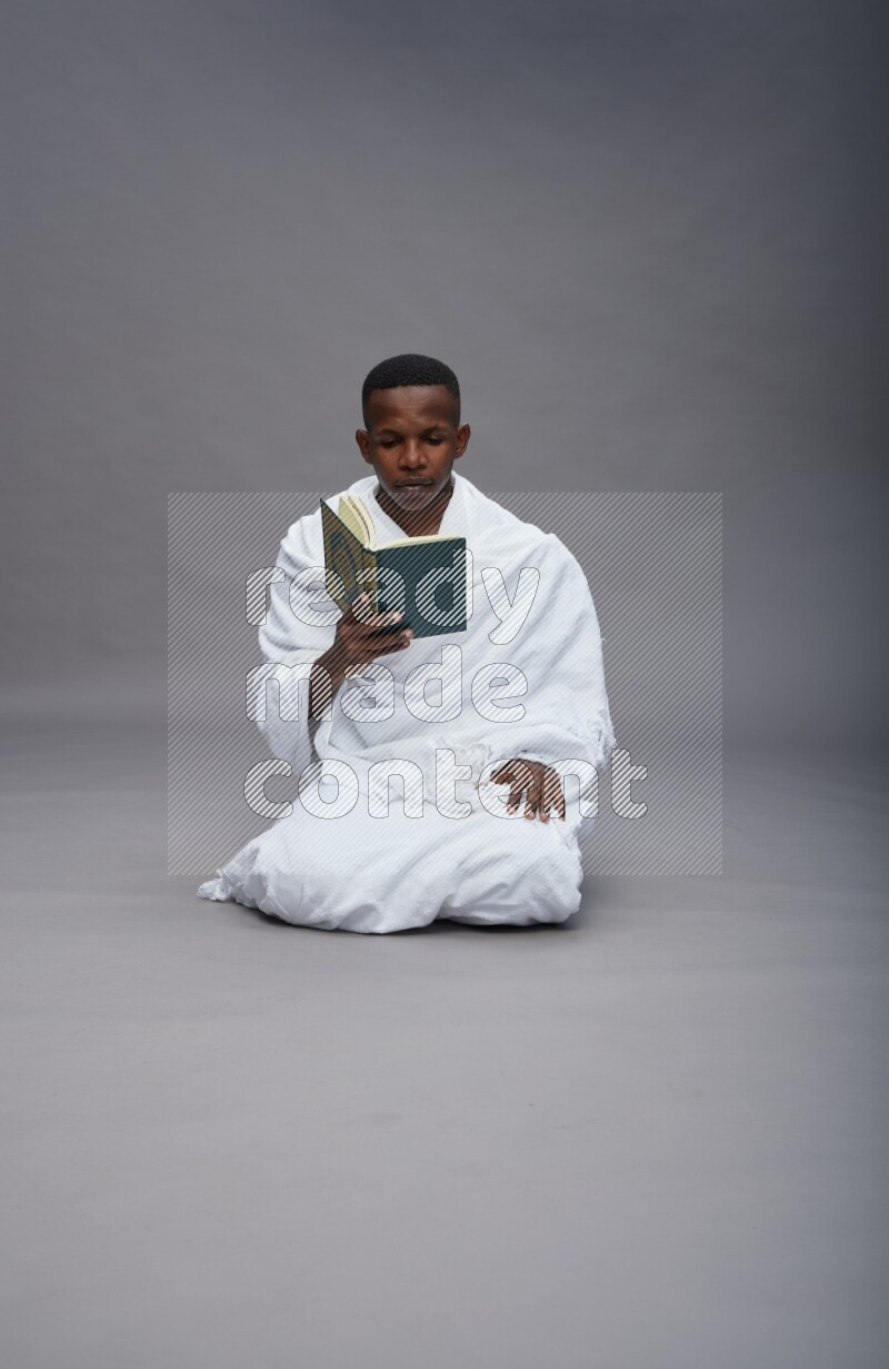 A man wearing Ehram sitting on floor reading quran on gray background