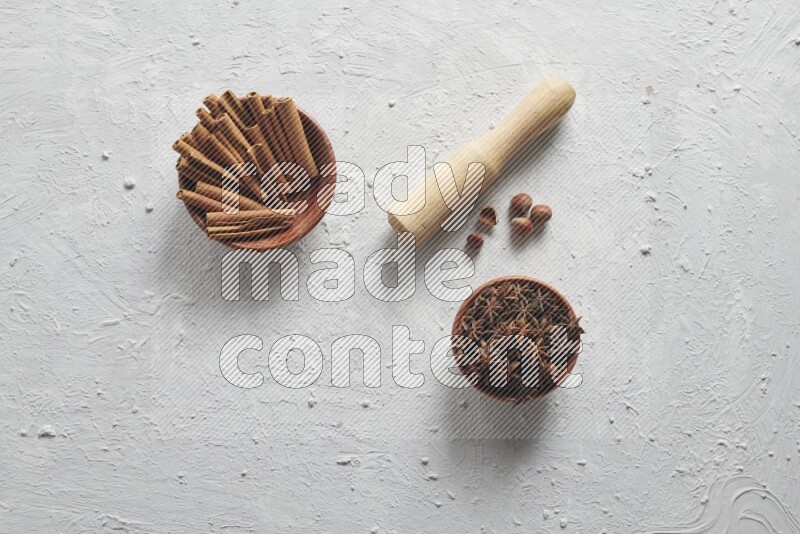 A wooden bowl full of cinnamon sticks and a wooden spoon with cinnamon powder are neatly placed beside a bowl of star anise on a textured white background