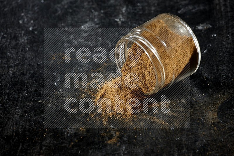 A flipped glass jar full of cumin powder and powder fell out on a textured black flooring