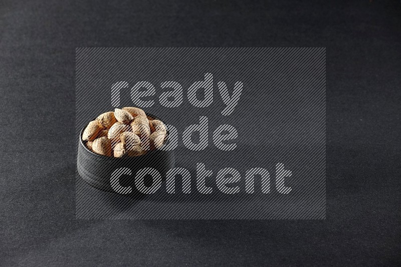A black pottery bowl full of almonds on a black background in different angles