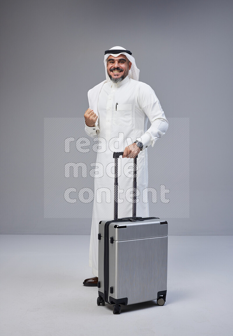 Saudi man wearing Thob and white Shomag standing holding Travel bag on Gray background