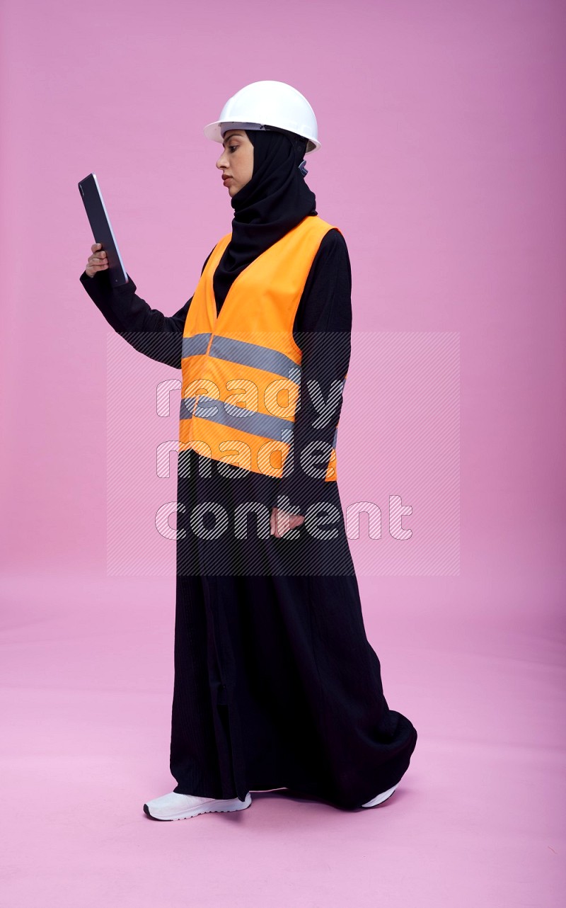 Saudi woman wearing Abaya with engineer vest and helmet standing working on tablet on pink background
