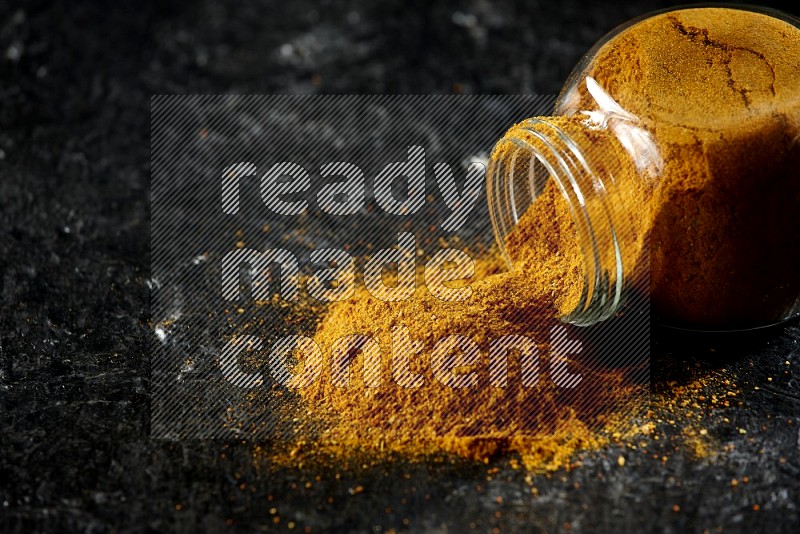 A flipped glass spice jar full of turmeric powder and powder spilled out of it on textured black flooring