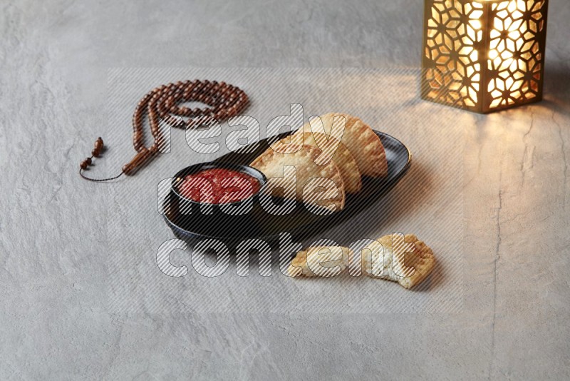 Three fried sambosas in an oval shaped black plate and a red sauce in a black round ramekin with a brown misbaha and a golden lantern on the side on a gray background