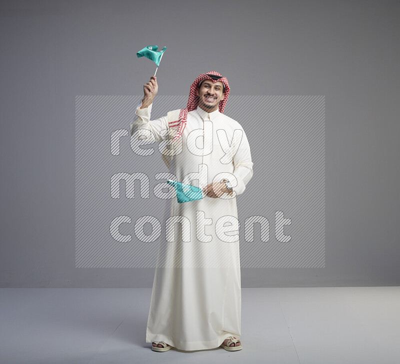 A Saudi man standing wearing thob and red shomag raising small Saudi flag on gray background