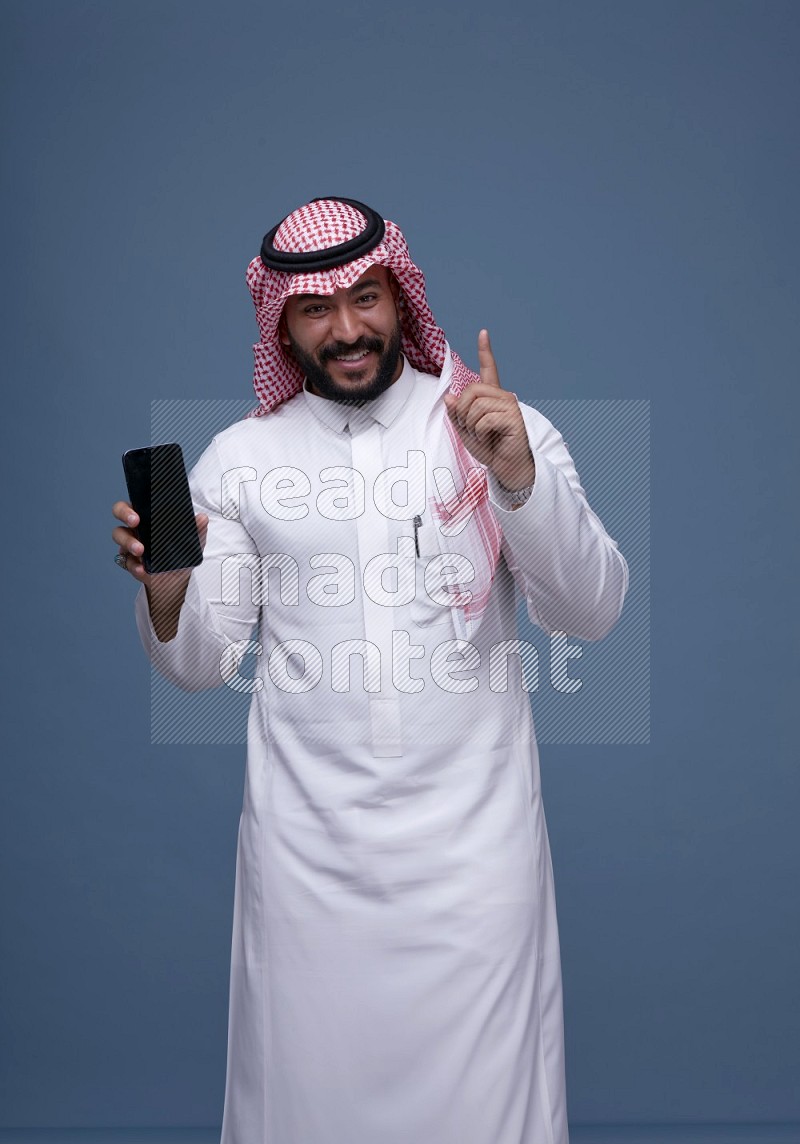 A man Showing his smartphone Screen on Blue Background wearing Saudi Thob