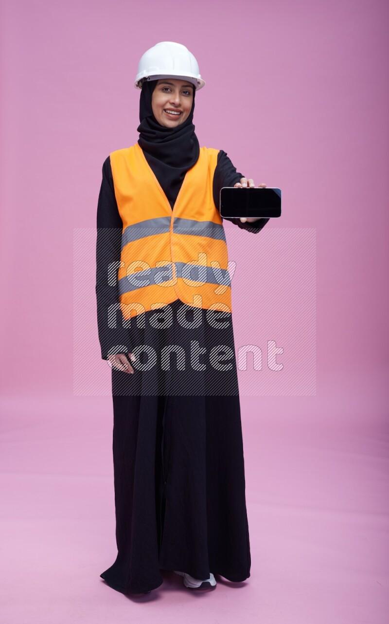 Saudi woman wearing Abaya with engineer vest and helmet standing showing phone to camera on pink background