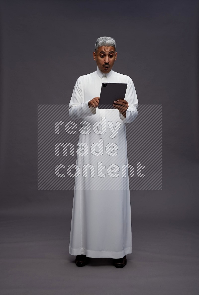 Saudi man wearing thob standing working on tablet on gray background