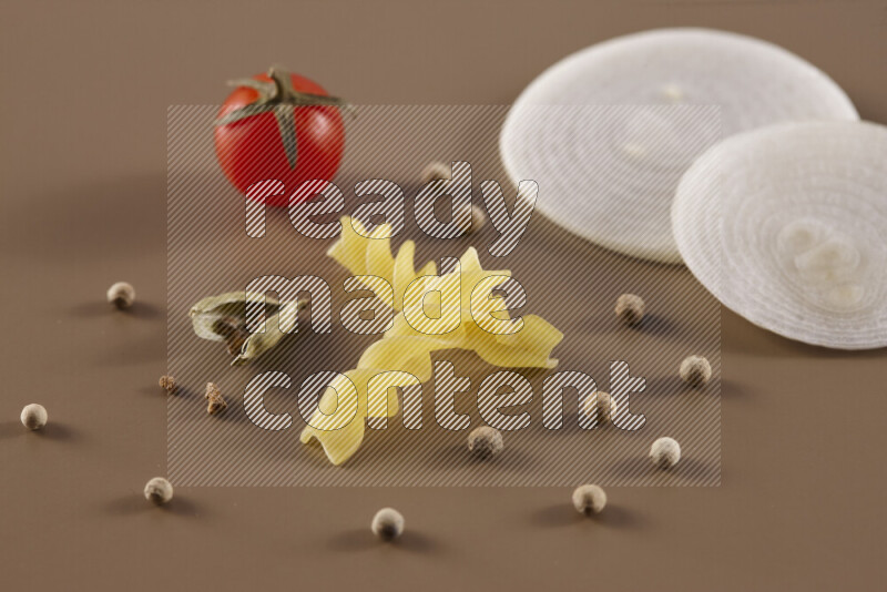 Raw pasta with different ingredients such as cherry tomatoes, garlic, onions, red chilis, black pepper, white pepper, bay laurel leaves, rosemary and cardamom on beige background