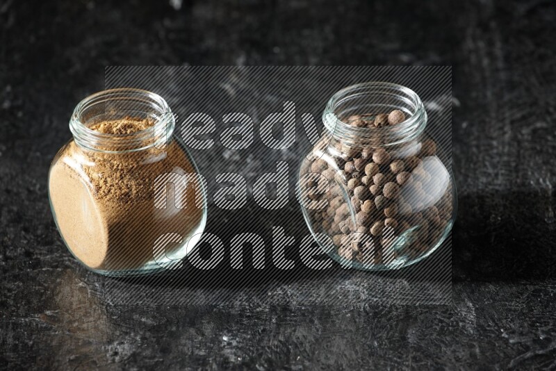 2 glass spice jars full of allspice powder and whole balls on a textured black flooring