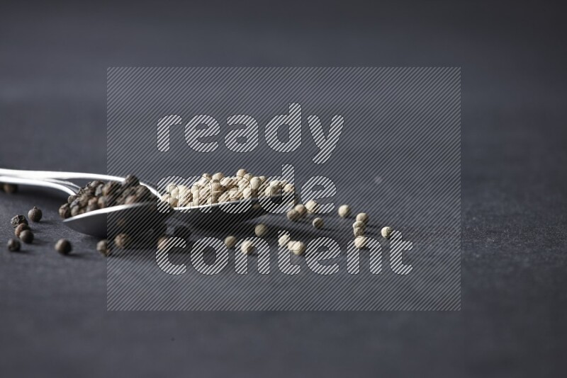 2 metal spoons full of black and white pepper beads with spreaded beads on black flooring