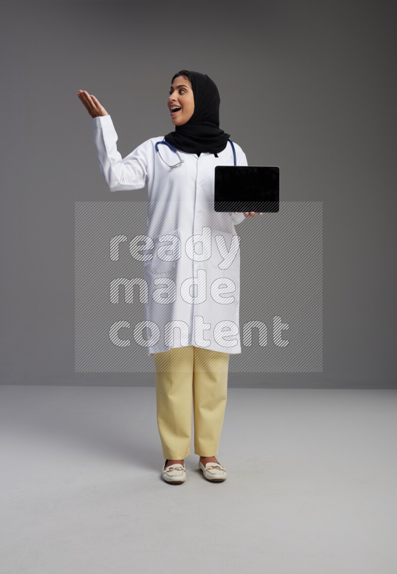 Saudi woman wearing lab coat with stethoscope standing showing tablet to camera with sign in the back on Gray background