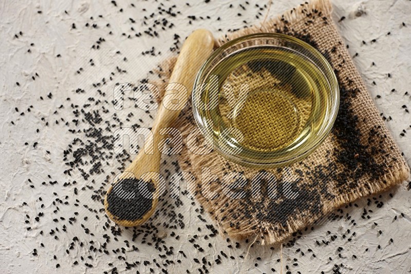 A glass bowl full of black seeds oil and wooden spoon full of black seeds with seeds spread on burlap fabric on a textured white flooring