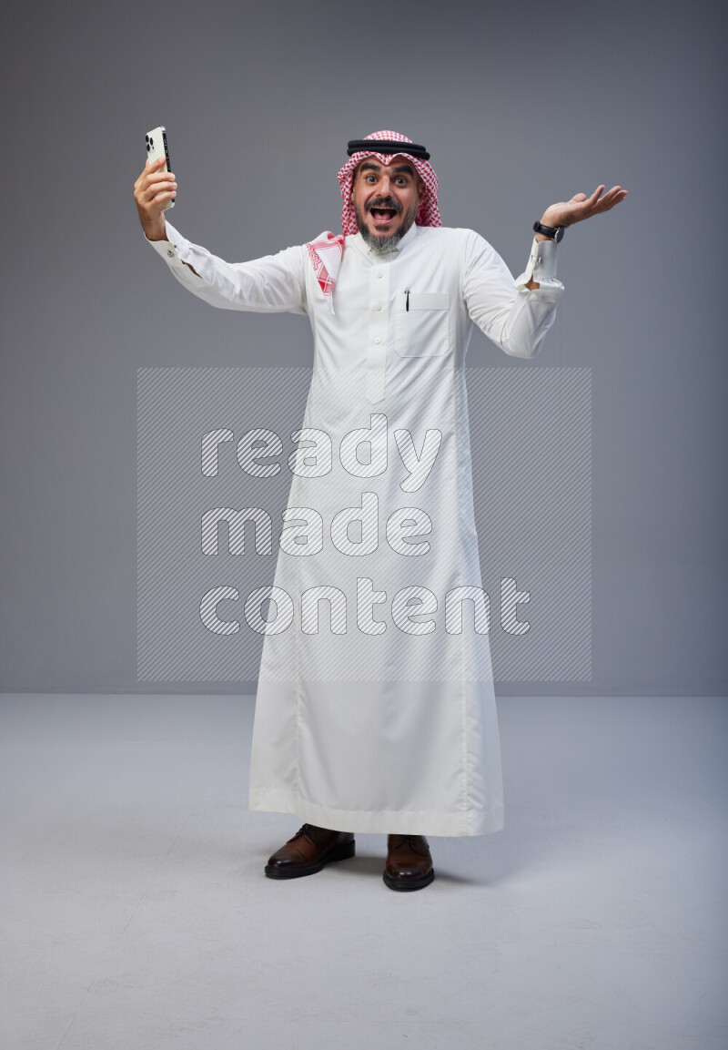 Saudi man Wearing Thob and red Shomag standing taking selfie on Gray background