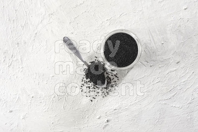 A glass jar and a metal spoon full of black seeds on a textured white flooring