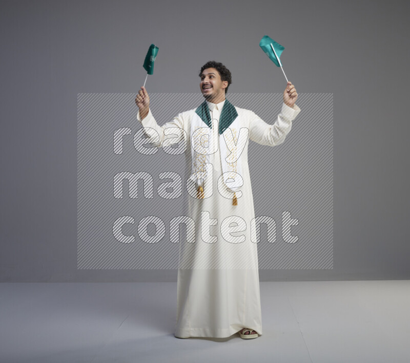A Saudi man standing wearing thob and saudi flag scarf and holding small saudi flag on gray background