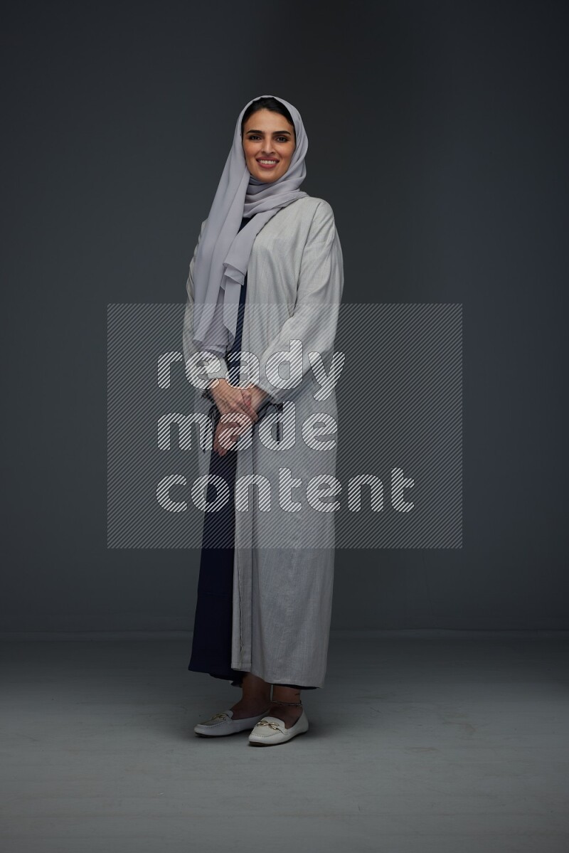 A Saudi woman wearing a light gray Abaya and head scarf standing and making multi poses eye level on a grey background