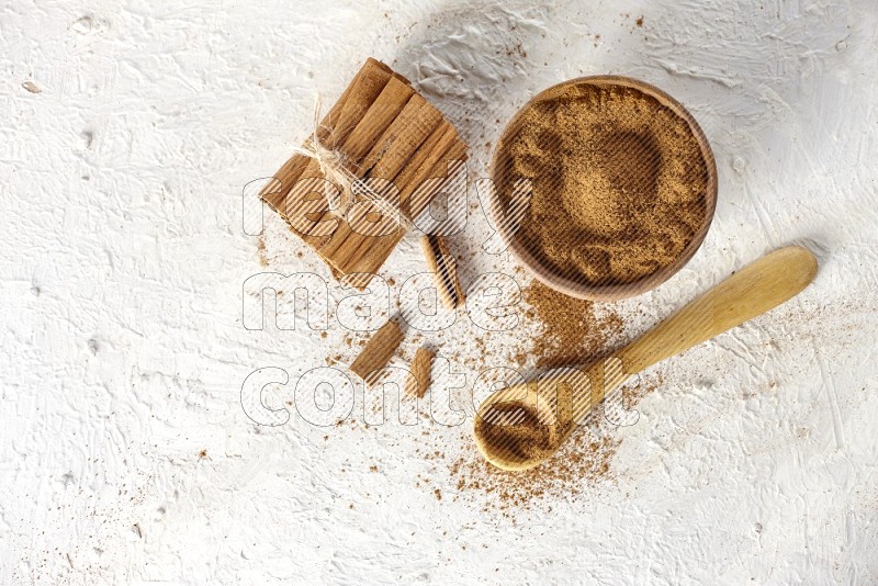 أعواد القرفة المربوطة بجانب وعاء خشبي مملوء ببودرة القرفة وملعقة خشبية مملوءة بالبودرة على خلفية بيضاء