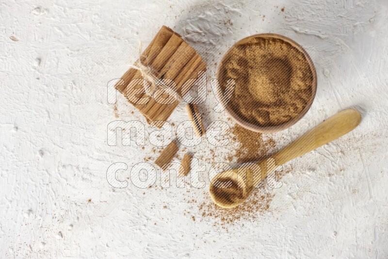 Cinnamon sticks stacked and bounded beside a wooden bowl full of cinnamon powder and a wooden spoon full of powder on white background