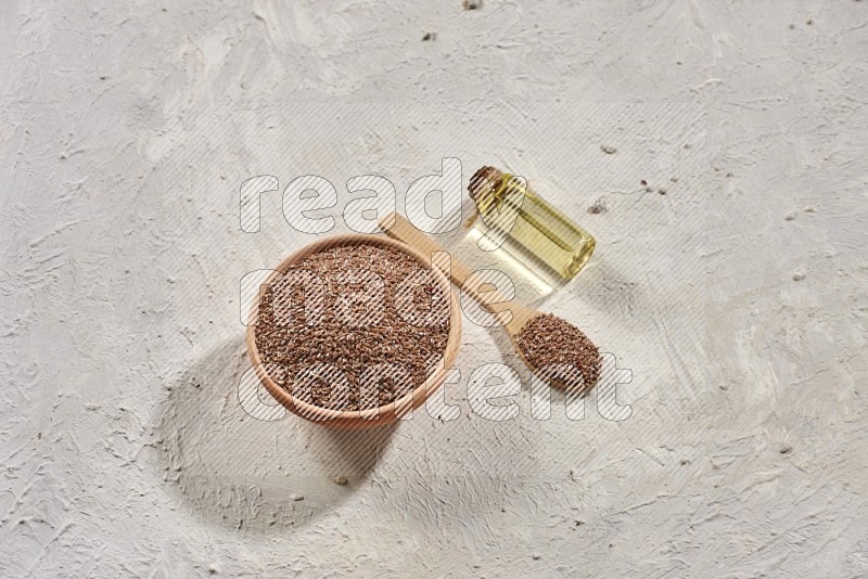 A wooden bowl and spoon full of flax and a bottle of flax oil on a textured white flooring in different angles