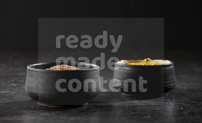 2 black pottery bowls full of mustard seeds and mustard paste on black flooring