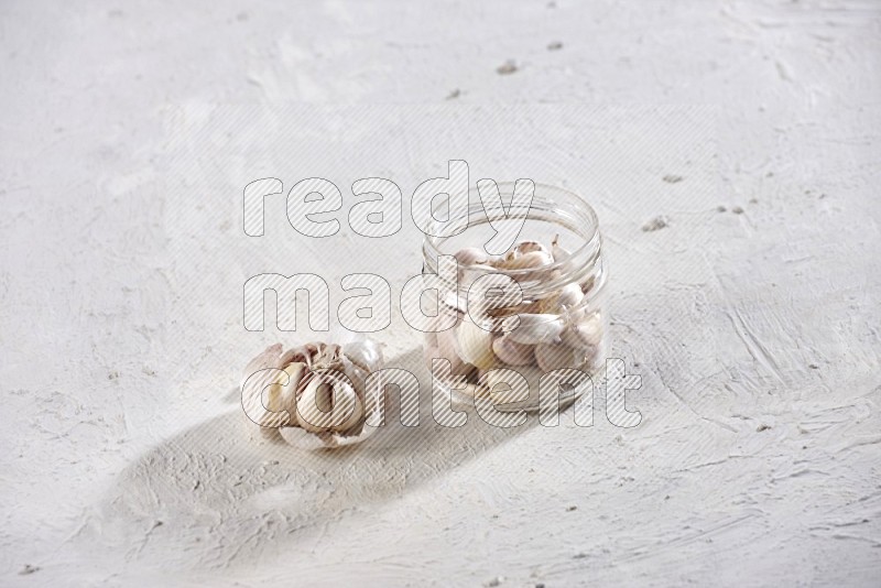 A glass jar full of garlic cloves and beside it a garlic bulb on a textured white flooring in different angles