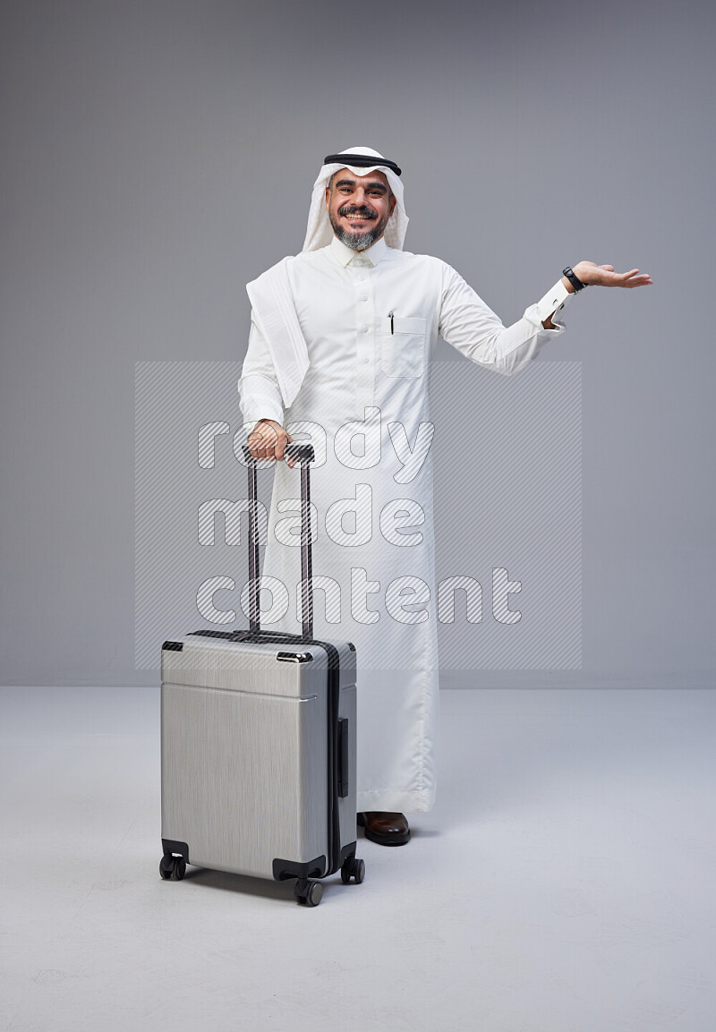 Saudi man wearing Thob and white Shomag standing holding Travel bag on Gray background