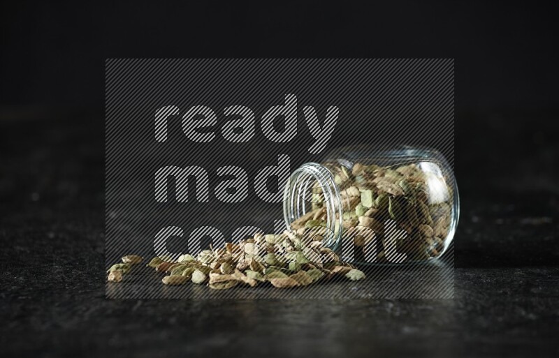 A flipped glass spice jar full of cardamom seeds on textured black flooring