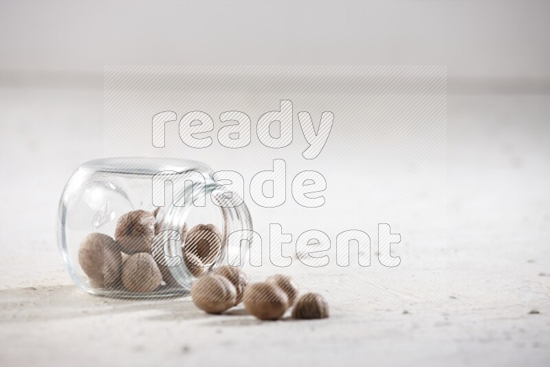 A glass spice jar full of whole nutmeg seeds flipped and the seeds spread out on a textured white flooring