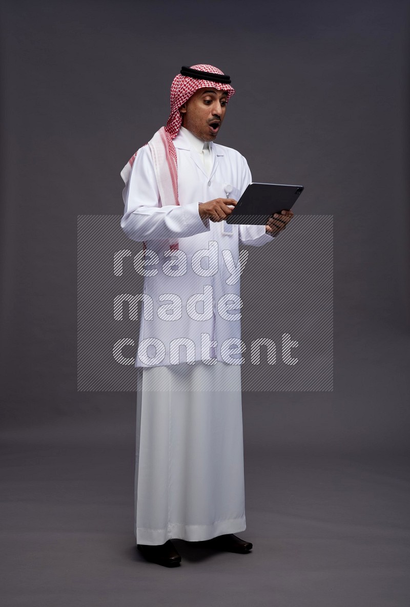 Saudi man wearing thob with lab coat and shomag with pocket employee badge standing working on tablet on gray background