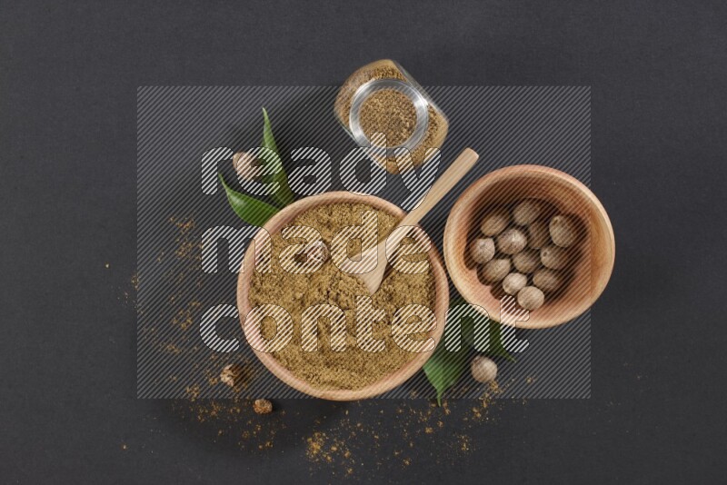A wooden bowl with a spoon in it and glass spice jar all, full of nutmeg powder and a wooden bowl full of whole nutmeg seeds on a black flooring