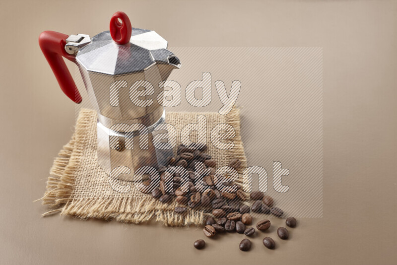 A moka pot with red handle surrounded by roasted coffee beans on beige background