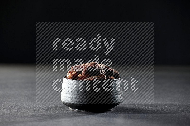 A black pottery bowl full of dried dates on a black background in different angles