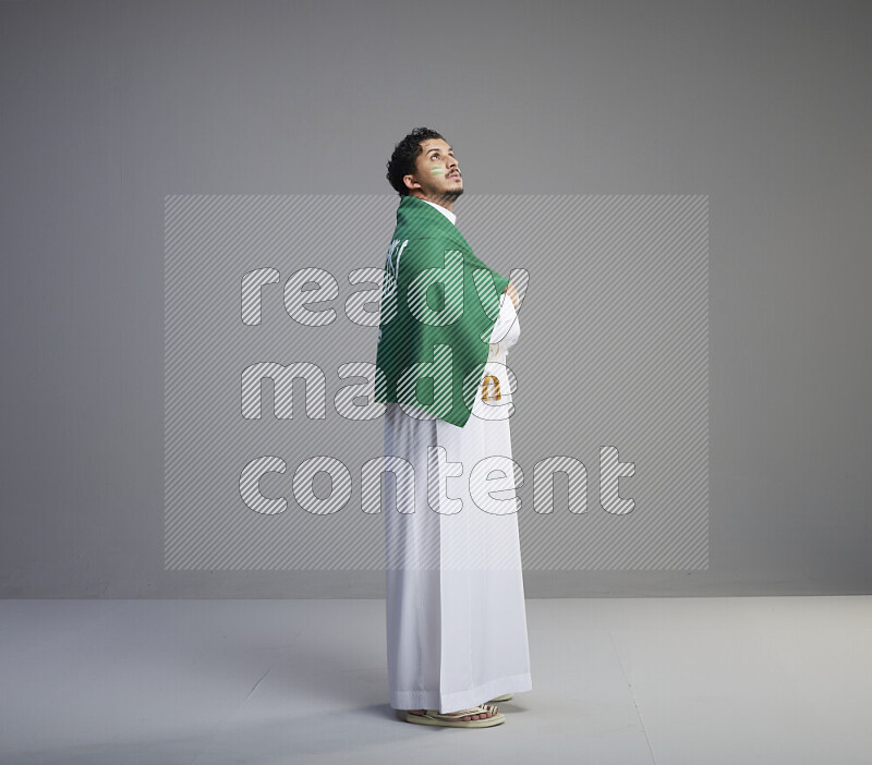 A Saudi man standing wearing thob and saudi flag scarf with face painting wrapping big Saudi flag on gray background