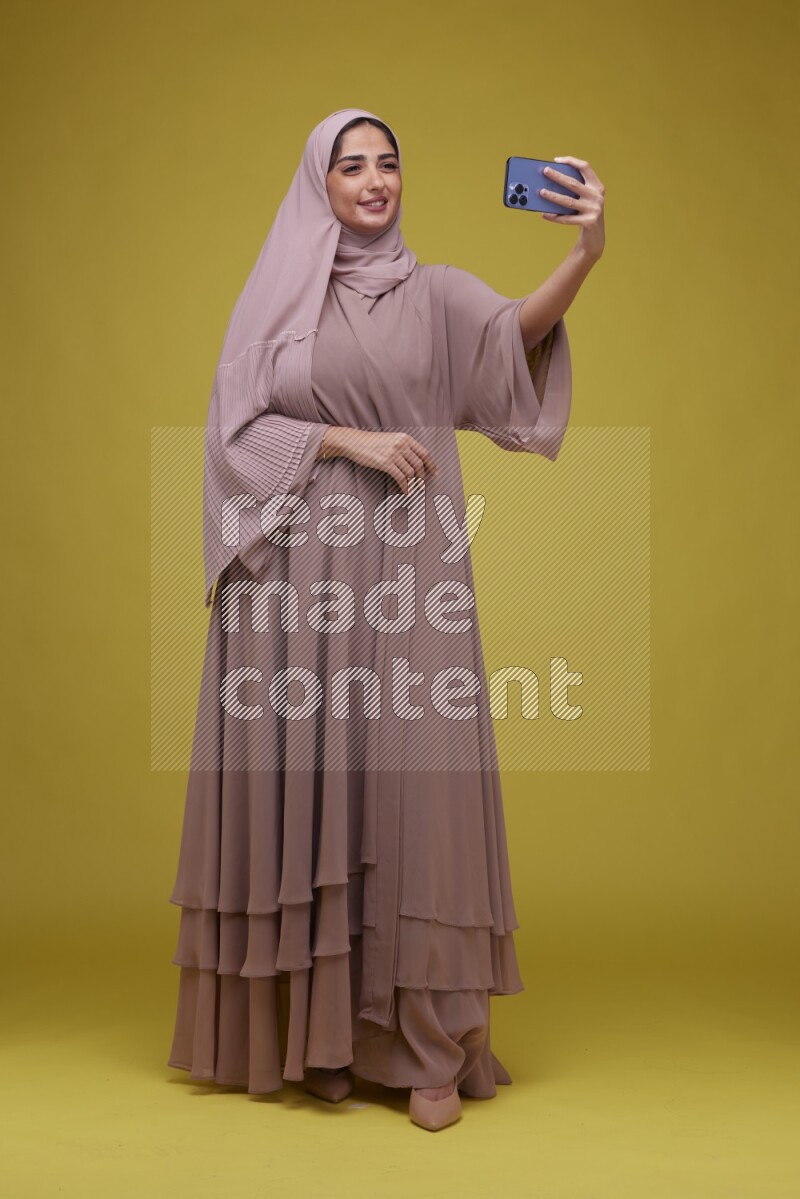 A woman taking a Selfie on a Yellow Background wearing Brown Abaya with Hijab