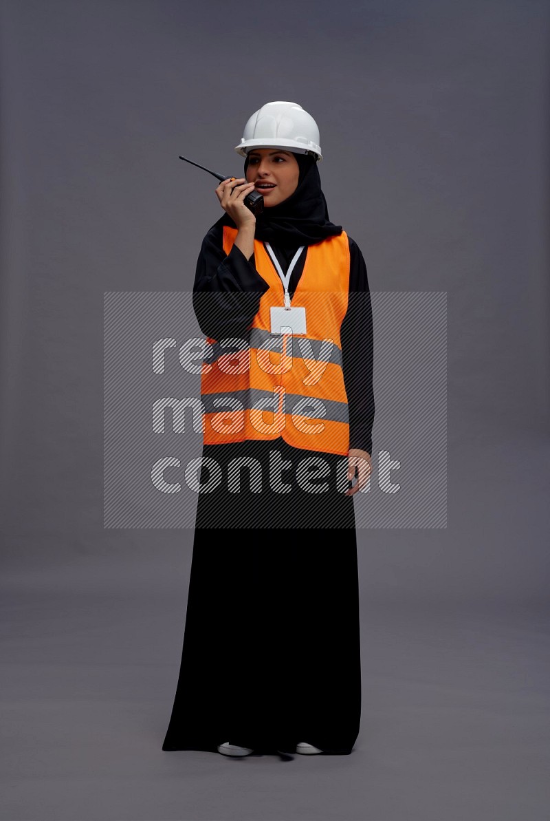 Saudi woman wearing Abaya with engineer vest with neck strap employee badge standing holding walkie-talkie on gray background