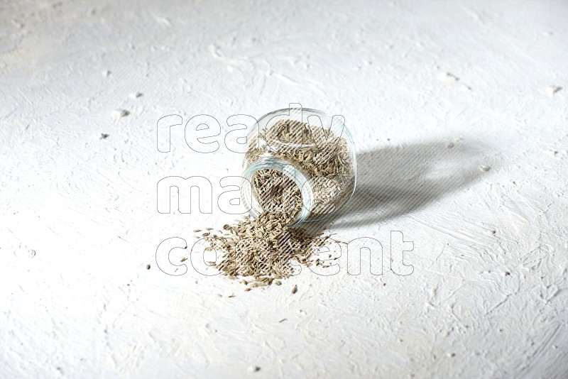 A flipped glass spice jar full of cumin seeds and the seeds spilled out on textured white flooring