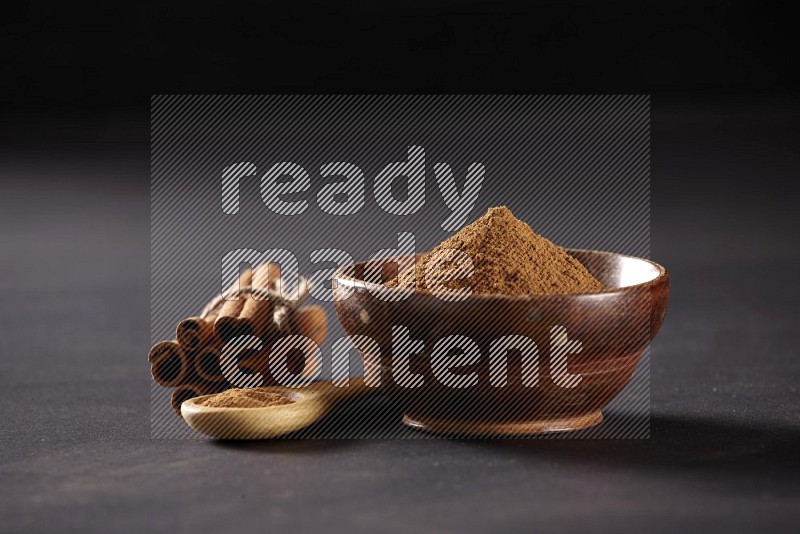 Cinnamon sticks stacked and bounded beside a wooden bowl full of cinnamon powder and a wooden spoon full of powder on black background