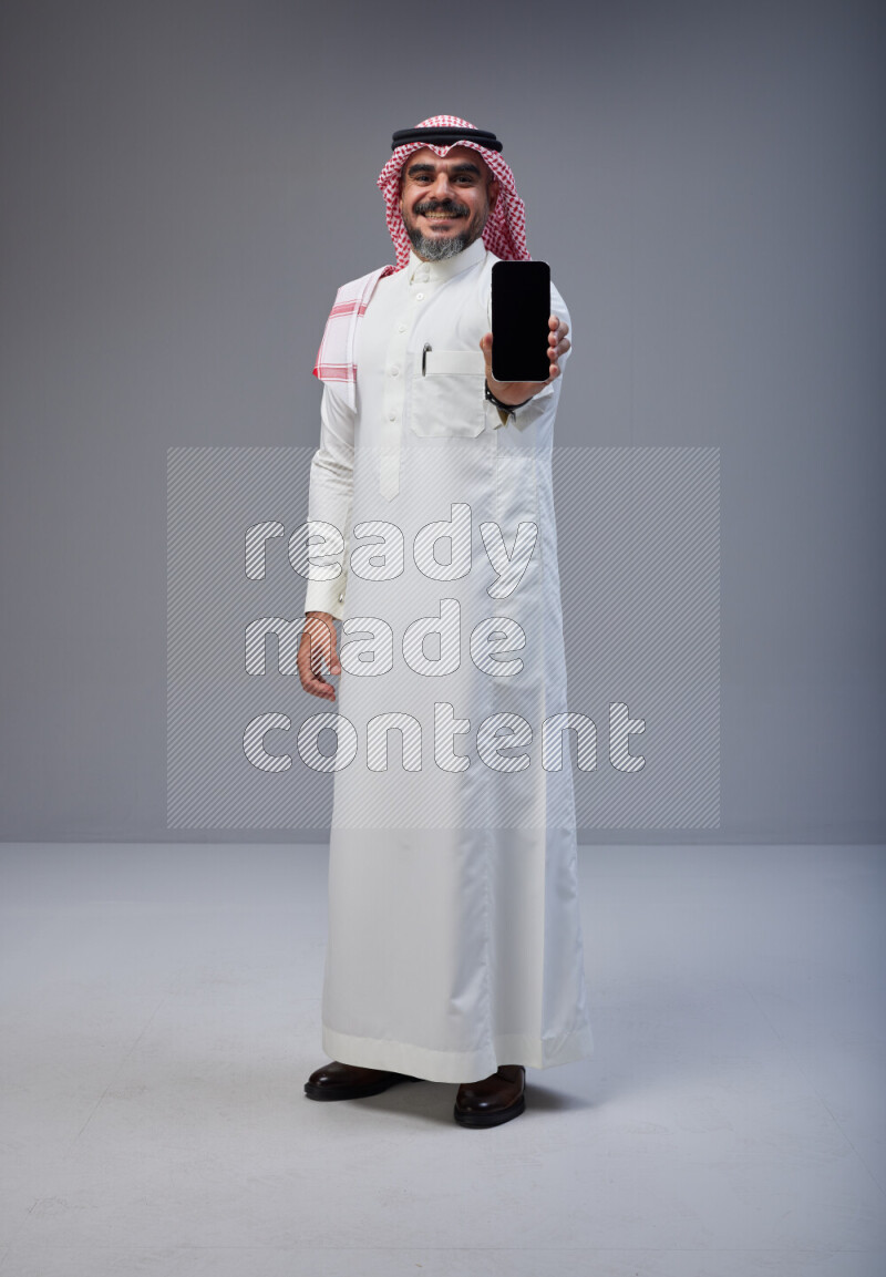 Saudi man Wearing Thob and red Shomag standing showing phone to camera on Gray background