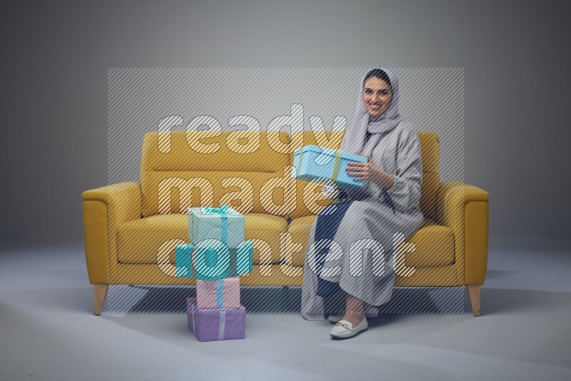 A Saudi woman wearing a light gray Abaya and head scarf sitting on a yellow sofa holding a gift box beside multi color gift boxes eye level on a grey background