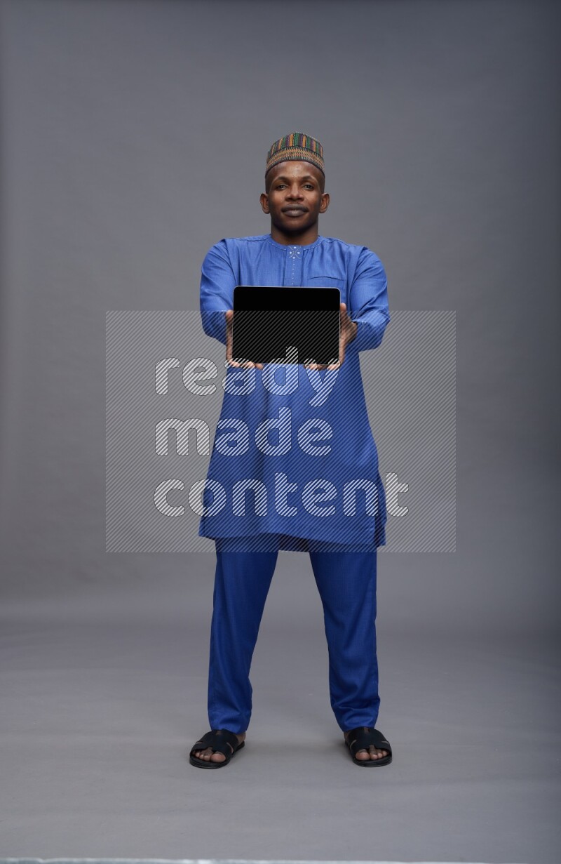 Man wearing Nigerian outfit standing showing tablet to camera on gray background