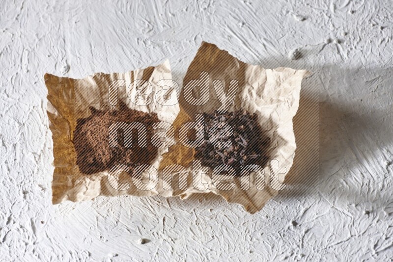 Cloves powder and cloves on 2 crumpled pieces of paper on a textured white flooring