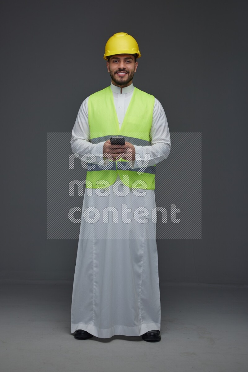 A Saudi man wearing Thobe with a yellow safety vest and white helmet standing and using his phone eye level on a gray background