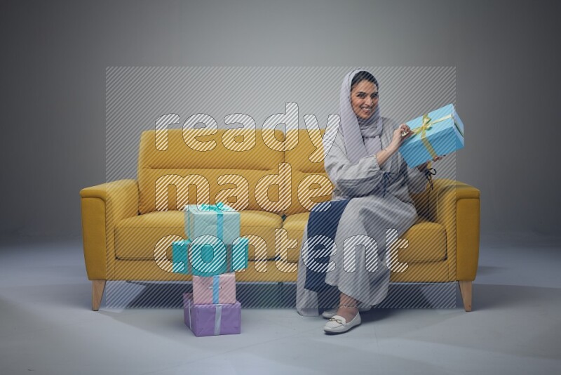 A Saudi woman wearing a light gray Abaya and head scarf sitting on a yellow sofa holding a gift box beside multi color gift boxes eye level on a grey background