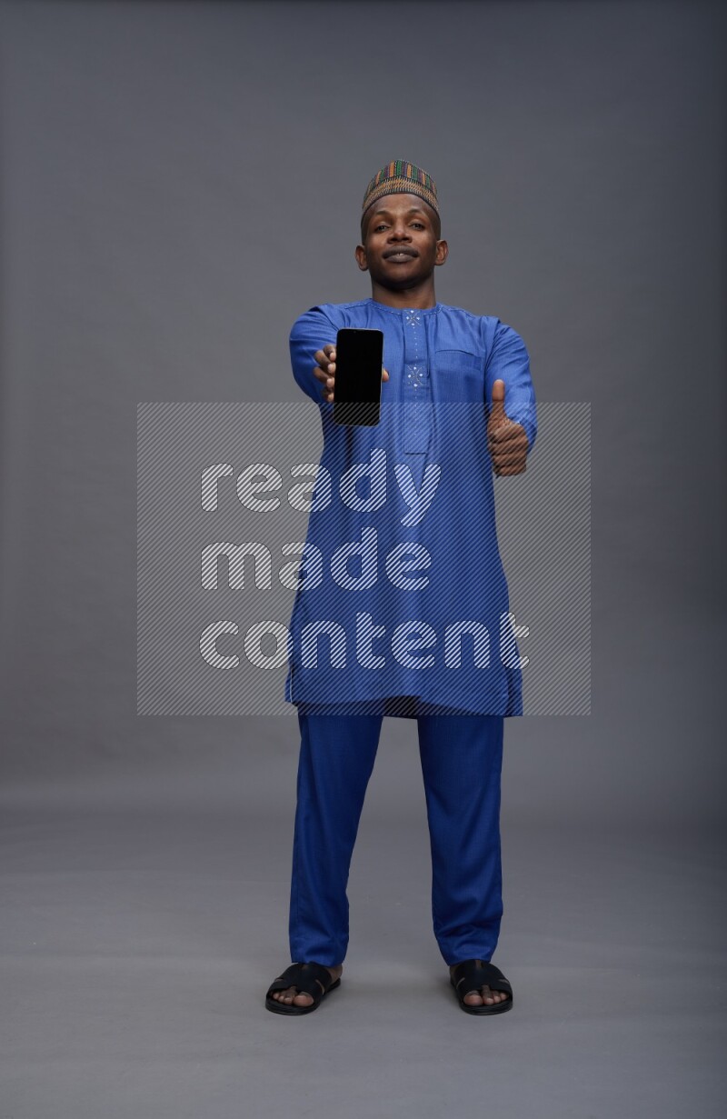 Man wearing Nigerian outfit standing showing phone to camera on gray background