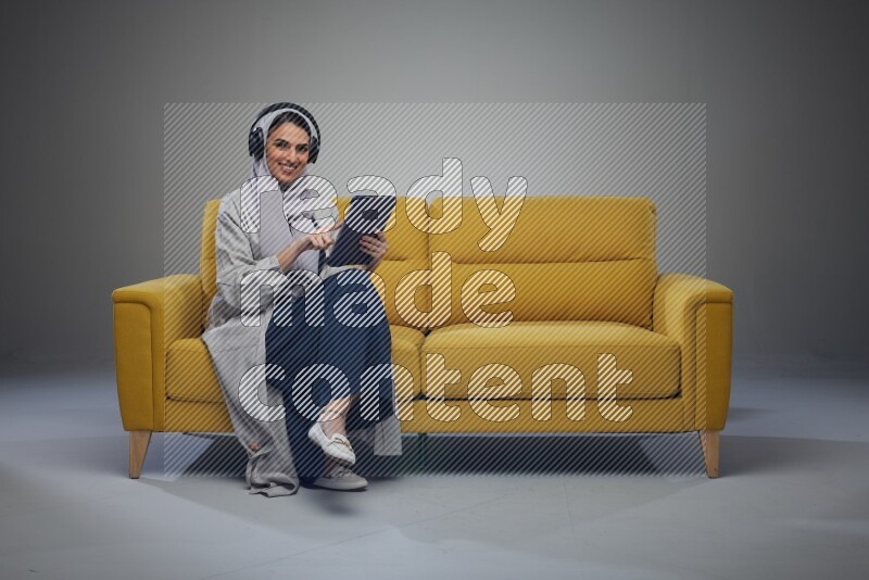 A Saudi woman wearing a light gray Abaya and head scarf sitting on a yellow sofa and using her phone while wearing headphones eye level on a grey background
